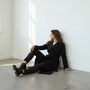 Woman in black wool coat seated against white wall in minimal studio setting